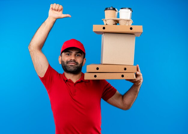 Repartidor barbudo en uniforme rojo y gorra sosteniendo cajas de cartón con una sonrisa en la cara mostrando los pulgares para arriba de pie sobre la pared azul