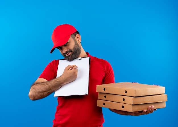 Repartidor barbudo en uniforme rojo y gorra sosteniendo cajas de pizza y portapapeles con páginas en blanco pidiendo firma sonriendo de pie sobre la pared azul