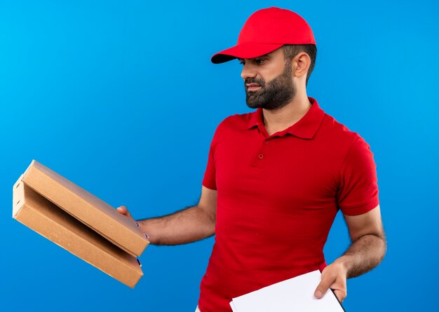 Repartidor barbudo en uniforme rojo y gorra sosteniendo cajas de pizza y portapapeles con páginas en blanco que parecen confundidos teniendo dudas sobre la pared azul