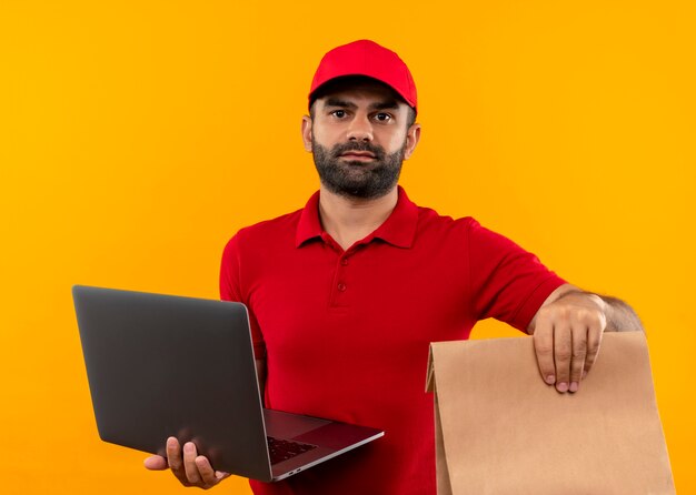 Repartidor barbudo en uniforme rojo y gorra sosteniendo paquete de papel y portátil con cara seria de pie sobre pared naranja