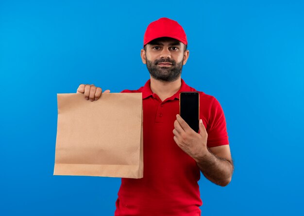 Repartidor barbudo en uniforme rojo y gorra sosteniendo el paquete de papel sonriendo mostrando smartphone parado sobre la pared azul