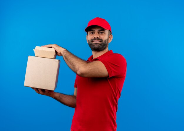 Repartidor barbudo en uniforme rojo y gorra sosteniendo paquetes de caja con sonrisa de confianza de pie sobre la pared azul