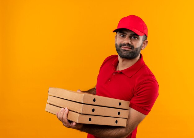 Repartidor barbudo en uniforme rojo y gorra sosteniendo una pila de cajas de pizza mirando confiado de pie sobre la pared naranja