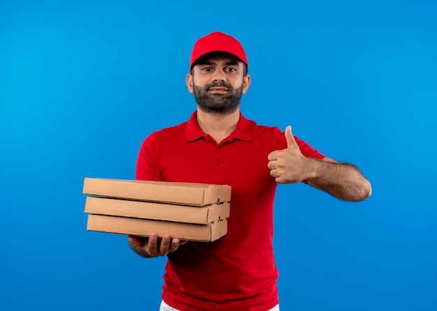 Repartidor barbudo en uniforme rojo y gorra sosteniendo una pila de cajas de pizza mostrando los pulgares para arriba sonriendo confiado de pie sobre la pared azul