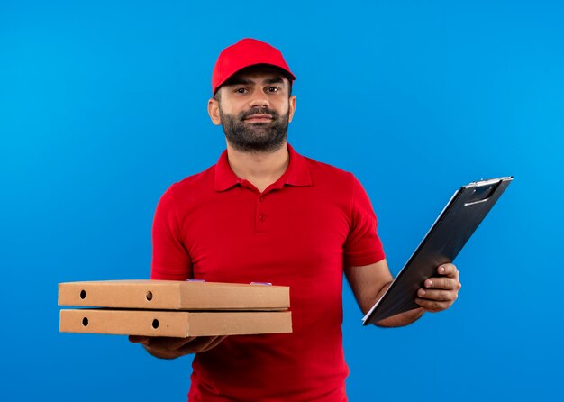 Repartidor barbudo en uniforme rojo y gorra sosteniendo una pila de cajas de pizza y portapapeles con expresión de confianza de pie sobre la pared azul