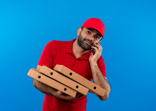 Repartidor barbudo en uniforme rojo y gorra sosteniendo una pila de cajas de pizza sonriendo mientras habla por teléfono móvil de pie sobre la pared azul