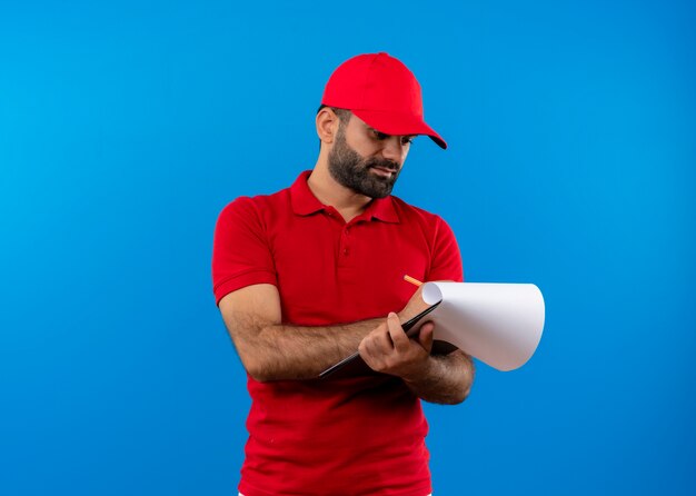 Repartidor barbudo en uniforme rojo y gorra sosteniendo portapapeles con páginas en blanco mirándolos con cara seria de pie sobre la pared azul