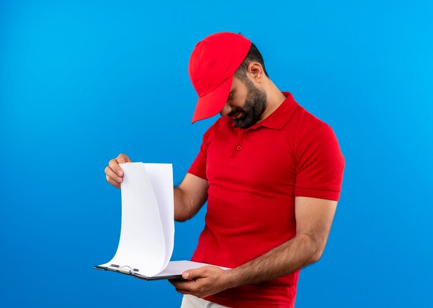 Repartidor barbudo en uniforme rojo y gorra sosteniendo portapapeles con páginas en blanco mirándolos con cara seria de pie sobre la pared azul