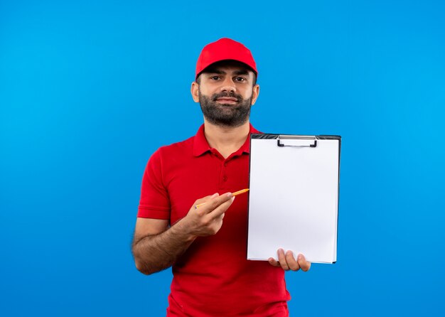 Repartidor barbudo en uniforme rojo y gorra sosteniendo el portapapeles con páginas en blanco pidiendo firma de pie sobre la pared azul