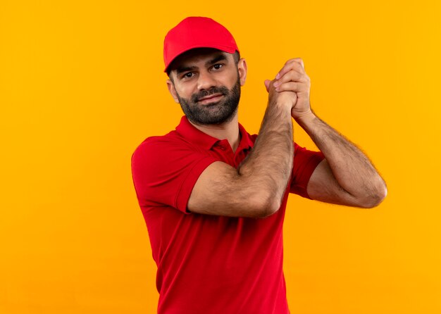 Repartidor barbudo en uniforme rojo y gorra tomados de la mano juntos haciendo gesto de trabajo en equipo sintiéndose agradecido de pie sobre la pared naranja