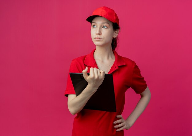 Repartidor bastante joven en uniforme rojo y gorra sosteniendo el portapapeles y mirando al lado con la mano en la cintura aislada sobre fondo carmesí con espacio de copia