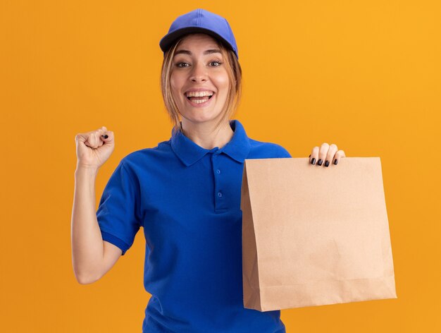 Repartidor bonita joven alegre en uniforme mantiene el puño y sostiene el paquete de papel en naranja