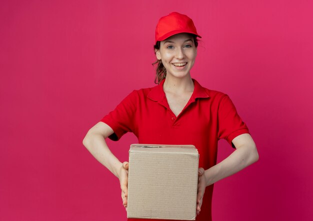 Repartidor bonita joven alegre en uniforme rojo y gorra con caja de cartón aislada sobre fondo carmesí con espacio de copia