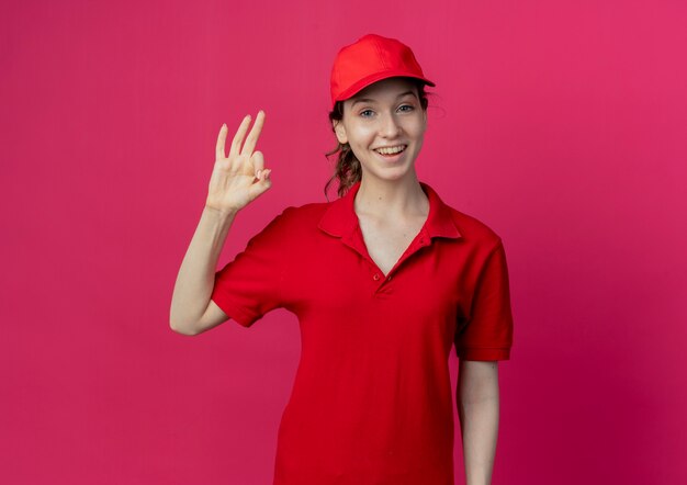 Repartidor bonita joven alegre en uniforme rojo y gorra haciendo bien firmar aislado sobre fondo carmesí con espacio de copia