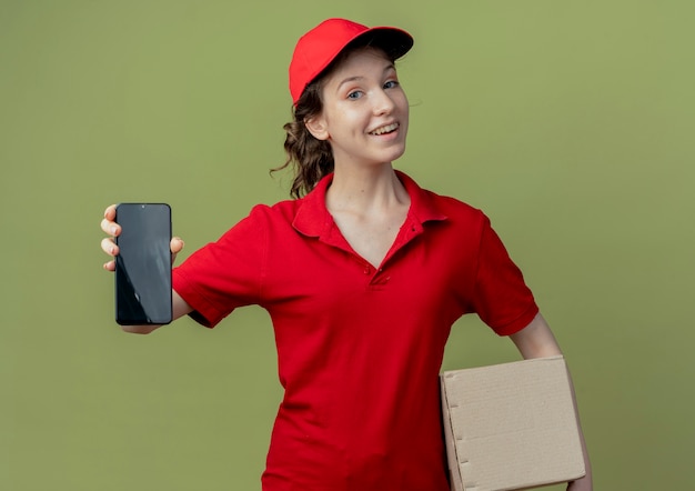 Repartidor bonita joven alegre en uniforme rojo y gorra sosteniendo la caja de cartón y estirando el teléfono móvil en la cámara aislada sobre fondo verde oliva