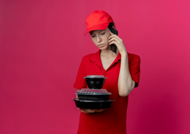 Repartidor bonita joven disgustada con uniforme rojo y gorra hablando por teléfono y sosteniendo y mirando contenedores de comida aislados sobre fondo carmesí con espacio de copia