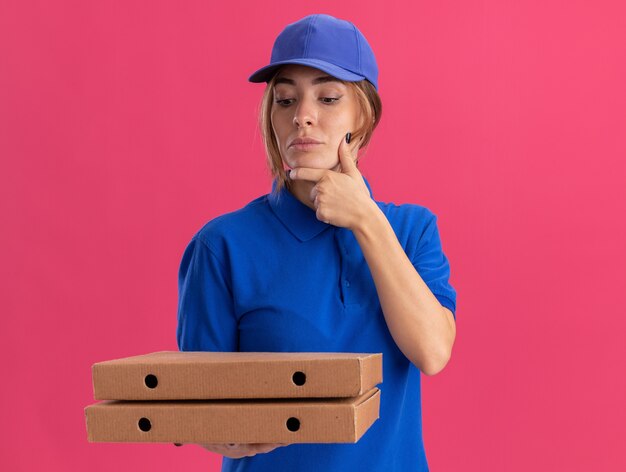 Repartidor bonita joven pensativa en uniforme pone la mano en la barbilla y mira cajas de pizza en rosa