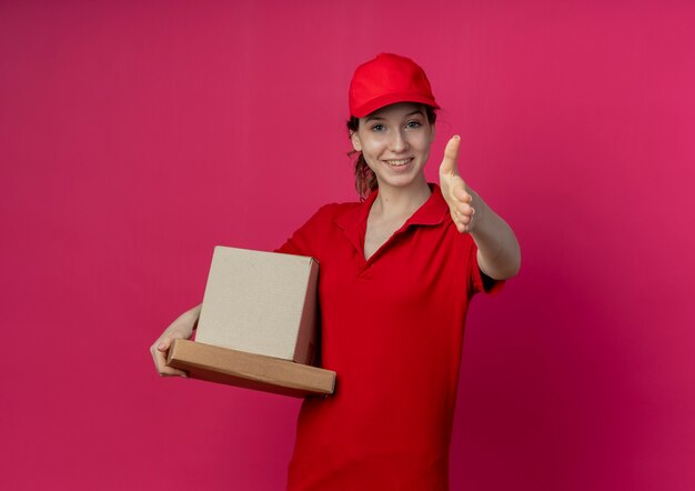 Repartidor bonita joven sonriente con uniforme rojo y gorra sosteniendo el paquete de pizza y caja de cartón extendiendo la mano hacia la cámara gesticulando hola aislado sobre fondo carmesí con espacio de copia