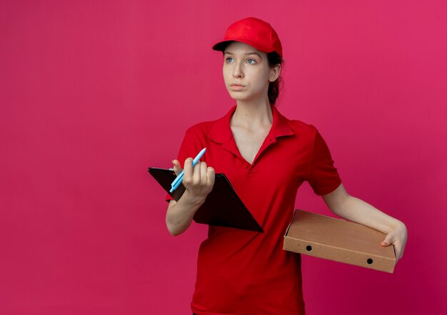 Repartidor bonita joven en uniforme rojo y gorra con bolígrafo de paquete de pizza y portapapeles mirando al lado aislado sobre fondo carmesí con espacio de copia