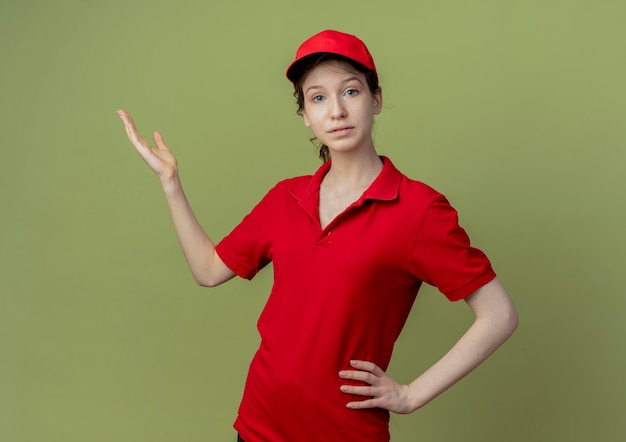 Foto gratuita repartidor bonita joven en uniforme rojo y gorra mirando a cámara poniendo la mano en la cintura y mostrando la mano vacía aislada sobre fondo verde oliva con espacio de copia