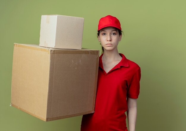 Repartidor bonita joven en uniforme rojo y gorra mirando a cámara y sosteniendo cajas de cartón aisladas sobre fondo verde oliva con espacio de copia