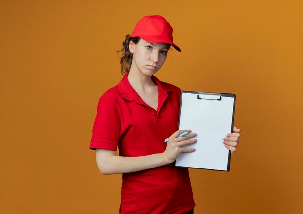 Repartidor bonita joven en uniforme rojo y gorra mirando a la cámara sosteniendo la pluma y mostrando el portapapeles en la cámara aislada sobre fondo naranja con espacio de copia