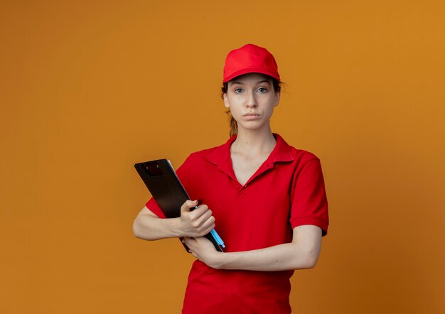 Repartidor bonita joven en uniforme rojo y gorra mirando a cámara sosteniendo portapapeles y bolígrafo aislado sobre fondo naranja con espacio de copia