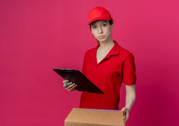 Repartidor bonita joven en uniforme rojo y gorra con paquete de pizza y portapapeles mirando a cámara aislada sobre fondo carmesí con espacio de copia