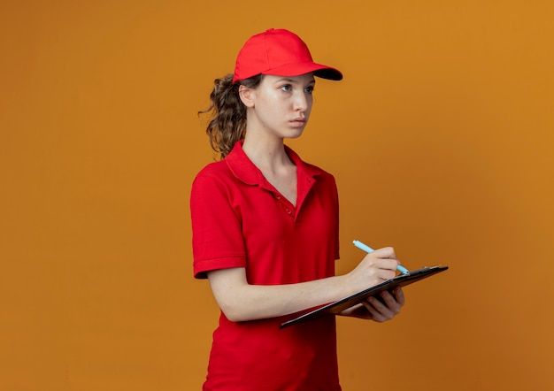 Repartidor bonita joven en uniforme rojo y gorra sosteniendo portapapeles y bolígrafo mirando directamente y preparándose para escribir aislado sobre fondo naranja con espacio de copia