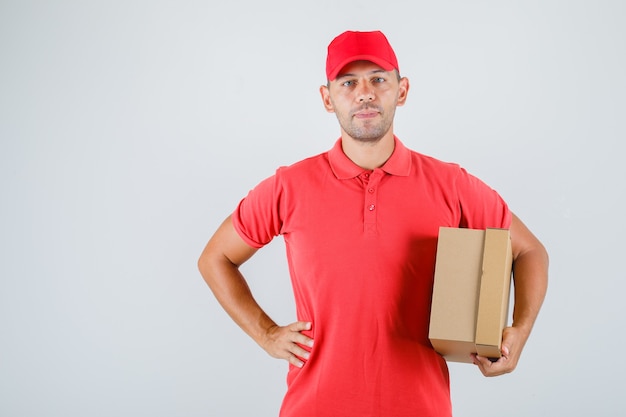 Repartidor con caja de cartón con la mano en la cintura en uniforme rojo, vista frontal.