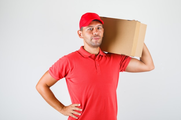 Repartidor con caja de cartón sobre su hombro en uniforme rojo y mirando positivo