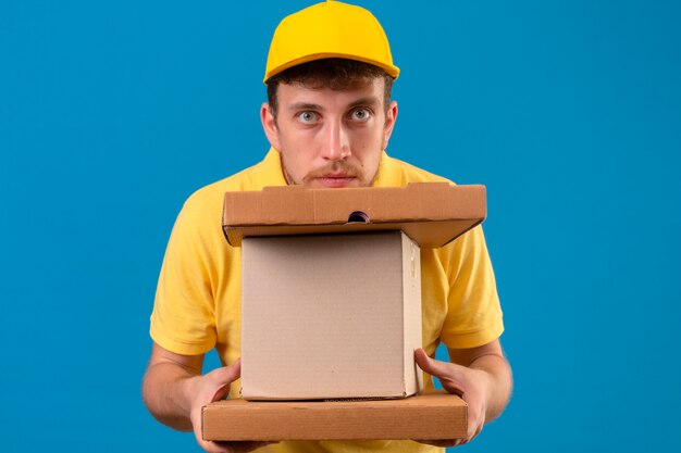 Repartidor en camisa polo amarilla y gorra con aspecto cansado y aburrido sosteniendo cajas de cartón en azul aislado