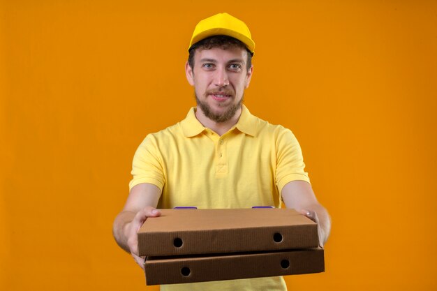 Repartidor en camisa polo amarilla y gorra estirando cajas de pizza dando a un cliente sonriendo amable en naranja aislado