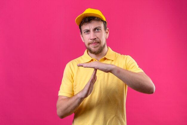 Repartidor en camisa polo amarilla y gorra mirando aburrido haciendo gesto de tiempo en rosa