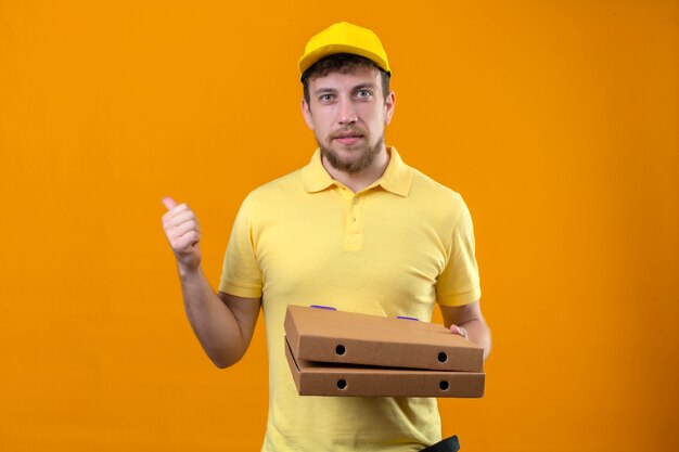 Repartidor en camisa polo amarilla y gorra mirando seguros mostrando Thumbs up de pie en naranja aislado
