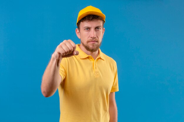 Repartidor en camisa polo amarilla y gorra mostrando el puño a la cámara con el ceño fruncido amenazando en azul aislado