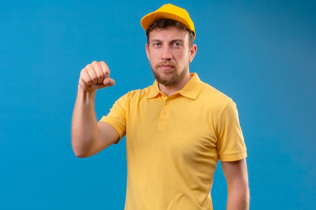 Repartidor en camisa polo amarilla y gorra mostrando el puño a la cámara con el ceño fruncido amenazando en azul aislado