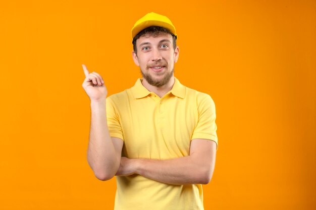 Repartidor en camisa polo amarilla y gorra sonriendo feliz apuntando con la mano y el dedo hacia el lado de pie en naranja aislado