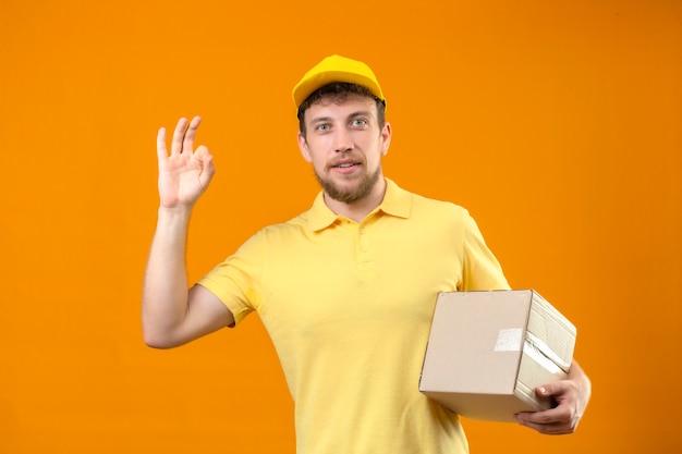 Repartidor en camisa polo amarilla y gorra sosteniendo una caja de cartón sonriendo amable haciendo bien firmar de pie en naranja aislado