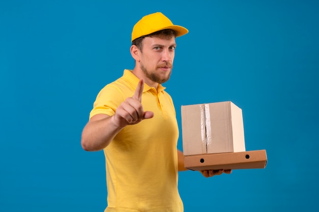 Repartidor en camisa polo amarilla y gorra sosteniendo cajas de cartón y apuntando a la cámara con el dedo con el ceño fruncido de pie en azul