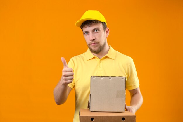 Repartidor en camisa polo amarilla y gorra sosteniendo cajas de cartón mirando confiado mostrando el pulgar hacia arriba de pie en naranja