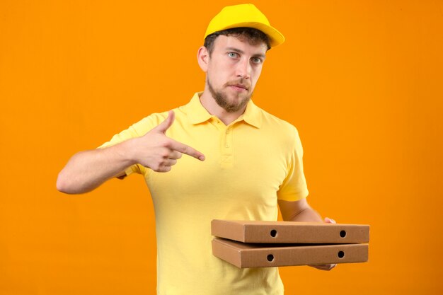 Repartidor en camisa polo amarilla y gorra sosteniendo cajas de pizza y apuntando con el dedo hacia ellos mirando confiado de pie en naranja aislado