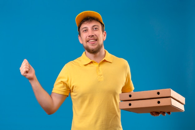 Repartidor en camisa polo amarilla y gorra sosteniendo cajas de pizza levantando el puño con cara feliz concepto ganador de pie sobre azul