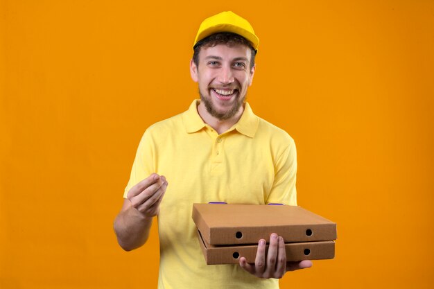 Repartidor en camisa polo amarilla y gorra sosteniendo cajas de pizza mirando a la cámara con una sonrisa en la cara haciendo gesto de dinero con la mano de pie en naranja