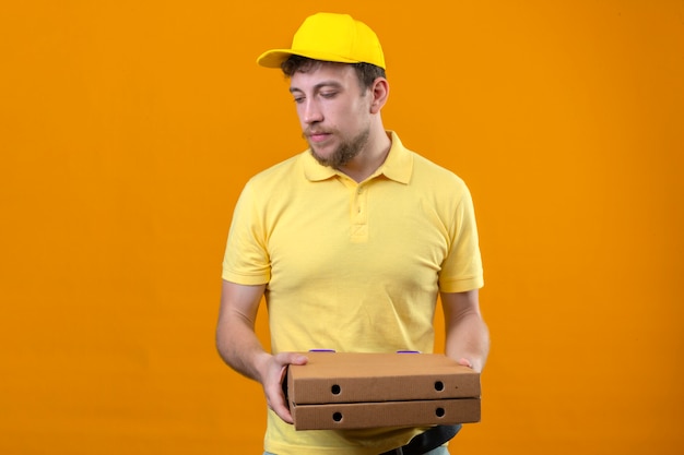 Repartidor en camisa polo amarilla y gorra sosteniendo cajas de pizza mirando a un lado con cara infeliz de pie en naranja