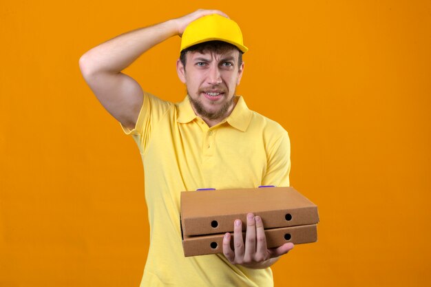 Repartidor con camisa polo amarilla y gorra sosteniendo cajas de pizza de pie con la mano en la cabeza por error recordar el error olvidó el concepto de mala memoria en naranja aislado
