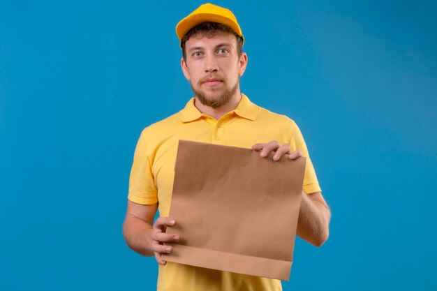 Repartidor en camisa polo amarilla y gorra sosteniendo el paquete de papel mirando a la cámara con cara seria de pie en azul aislado