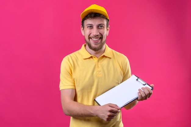 Repartidor en camisa polo amarilla y gorra sosteniendo el portapapeles en las manos mirando a la cámara con cara feliz de pie en rosa