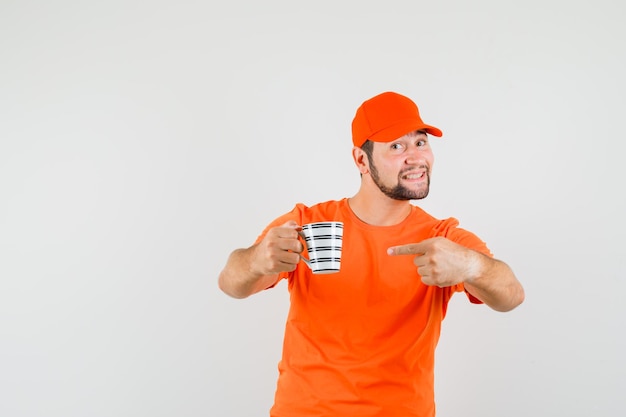 Repartidor en camiseta naranja, gorra apuntando a una taza de bebida y mirando optimista, vista frontal.