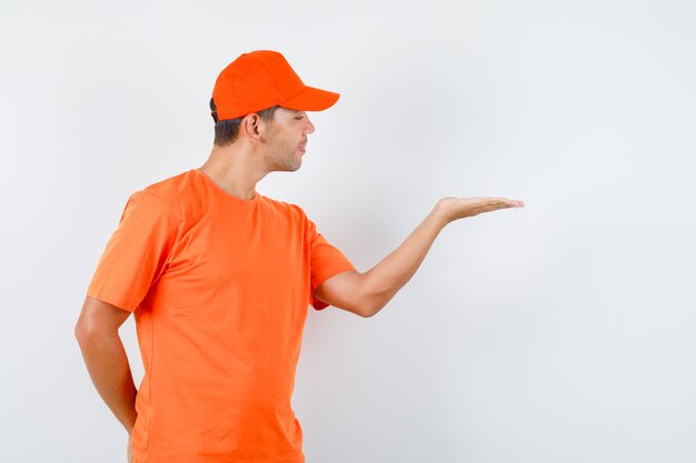 Repartidor en camiseta naranja y gorra extendiendo la palma levantada, ocultando la otra mano y mirando bromista, vista frontal.